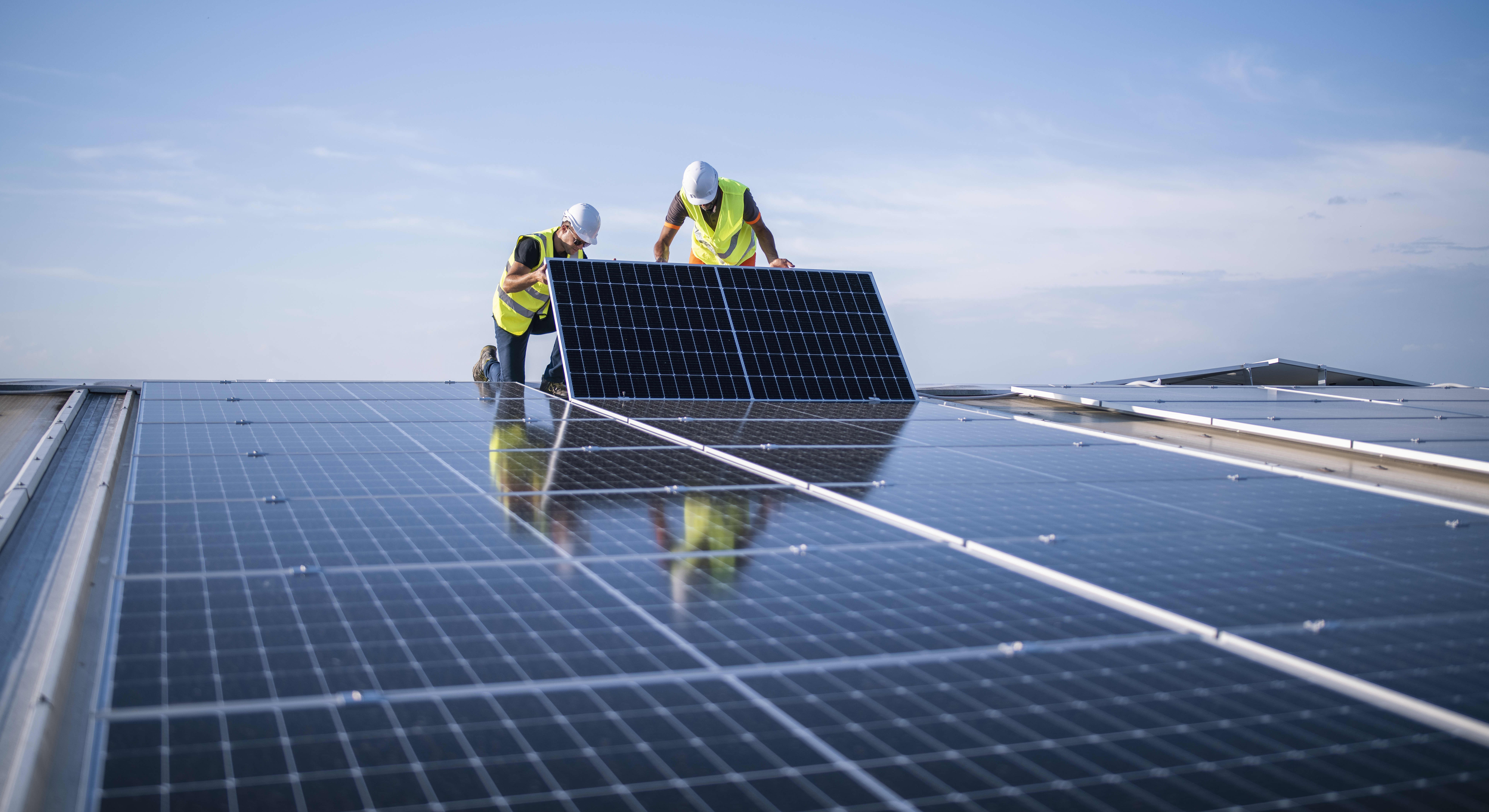 Zwei Arbeiter in gelben Warnwesten und weißen Helmen installieren ein Solarmodul auf einem Dach