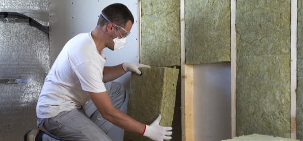 Handwerker mit Sichtschutzbrille und Atemmaske legt Steinwolle in Holzrahmen in einer Hauswand ein