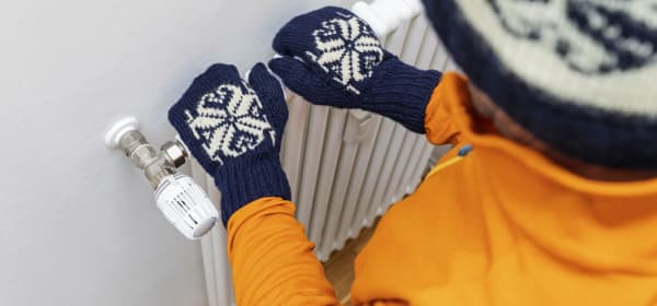 Kind in orangener Jacke und blauweißen Handschuhen und Mütze fasst auf Heizkörper