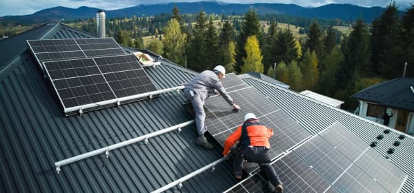 Zwei Installateure errichten eine Solaranlage auf einem Wohnhaus vor schöner Waldkulisse
