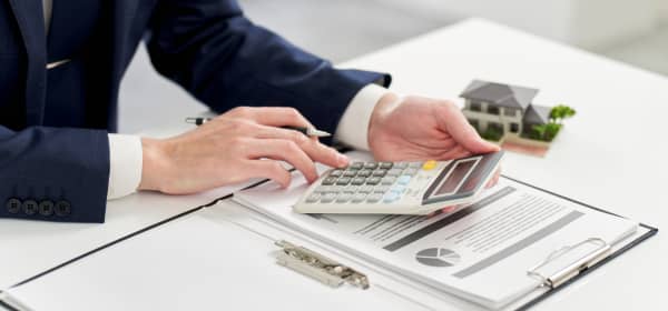 Mann in Anzug sitzt an einem Tisch mit Unterlagen und hält einen Taschenrechner in der Hand