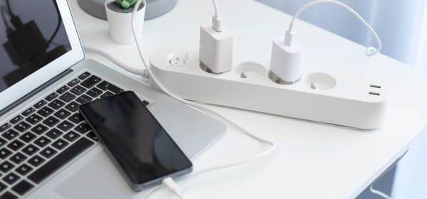 Weiße Steckerleiste mit eingesteckten Kabeln, die zu einem Laptop und einem Smartphone führen, die auf einem weißen Tisch liegen.