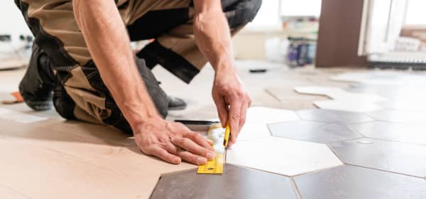 Mann in Arbeitskleidung kniet auf dem Boden und misst die Kante einer sechseckigen Bodenfliese zum Verlegen ab