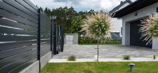 Modernes Wohnhaus mit gepflasterter Einfahrt und grüner Wiese. Das Grundstück wird durch einen hohen Zaun begrenzt.