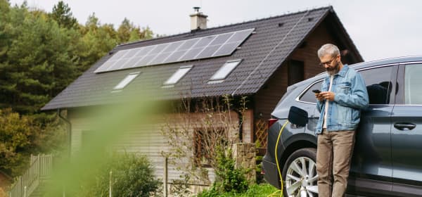 Mann steht mit Smartphone in der Hand an seinem Elektroauto, das gerade lädt, vor einem Haus mit Solarmodulen auf dem Dach