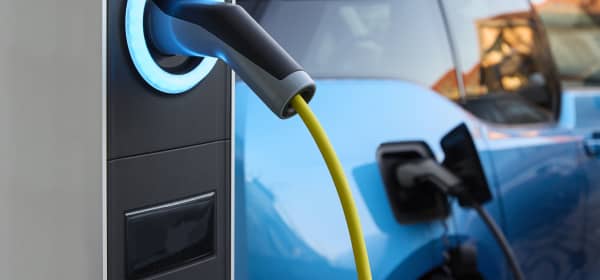 Ladesäule mit angeschlossenem Ladestecker und E-Auto im Hintergrund