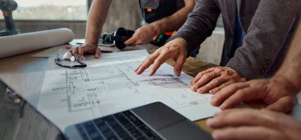 Drei Personen stehen an einem Tisch, auf dem Baupläne, ein Bohrer und ein Laptop liegen