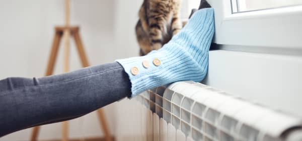 Füße einer jungen Frau auf einem Heizkörper unter einem gekippten Fenster, daneben sitzt eine Katze auf der Heizung und schaut nach draußen