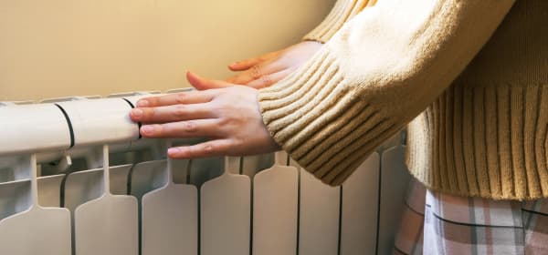 Frau in beigem Pullover und kariertem Rock wärmt ihre Hände an einem Heizkörper