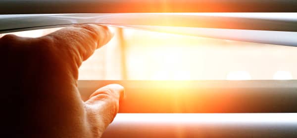 Hand einer Person, die die Lamellen einer Jalousie öffnet, dabei scheint die Sonne scheint durch das Fenster ins Hausinnere.