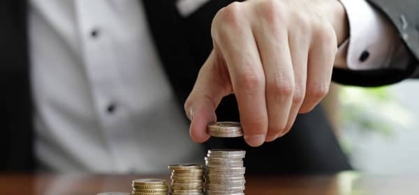 Mann in Hemd und Sakko stapelt Münzen auf ansteigend hohe Stapel auf einem Tisch