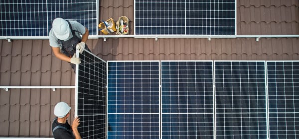 Ansicht von oben auf ein braun gedecktes Dach, auf dem zwei Arbeiter Solarmodule installieren