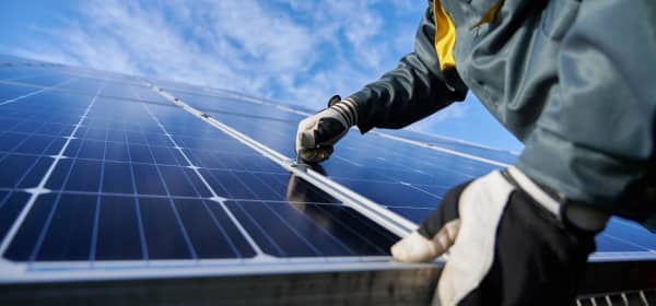 Nahaufnahme des Technikers in Arbeitshandschuhen, der eine Photovoltaikanlage unter blauem Himmel mit Wolken installiert