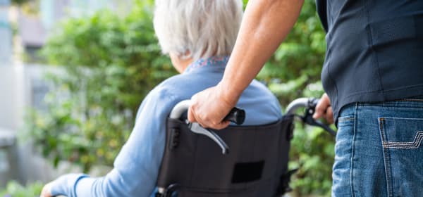 Rückansicht eines jüngeren Mannes, der eine Seniorin in einem Rollstuhl schiebt