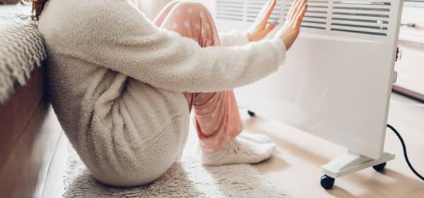 Frau sitzt auf dem Fußboden, lehnt sich dabei an einem Sofa an und streckt die Hände in Richtung einer Elektroheizung, die vor ihr steht