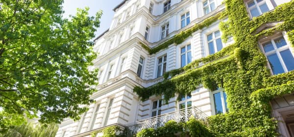 Blick von der Straße nach oben auf die weiße, teilweise mit Grünpflanzen bewachsene Fassade eines Mehrfamilienhauses