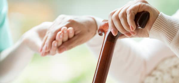 Nahaufnahme der Hände zweier Personen. Eine jüngere Person hält die Hand der älteren, die in der anderen Hand einen Gehstock hat