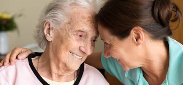 Frau im mittleren Alter lehnt ihre Stirn an die einer Seniorin, um deren Schultern sie einen Arm gelegt hat