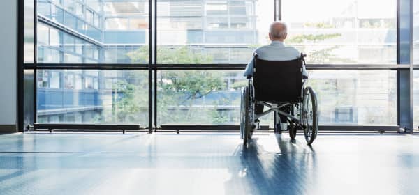 Senior im Rollstuhl blickt aus den bodentiefen Fenstern eines Gebäudes nach draußen