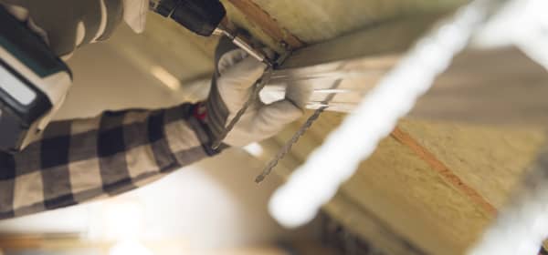 Nahaufnahme der Hände eines Arbeiters, der mit einem Akkuschrauber Bauteile innen an einer Dachschräge befestigt