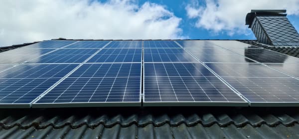 Blick von unten auf ein grau gedecktes Dach mit Schornstein und Solarmodulen bei blauem, leicht bewölktem Himmel