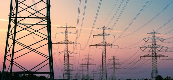 Mehrere Hochspannungsmasten in ländlicher Umgebung, die sich vor einem bewölkten Himmel bei Sonnenuntergang abzeichnen