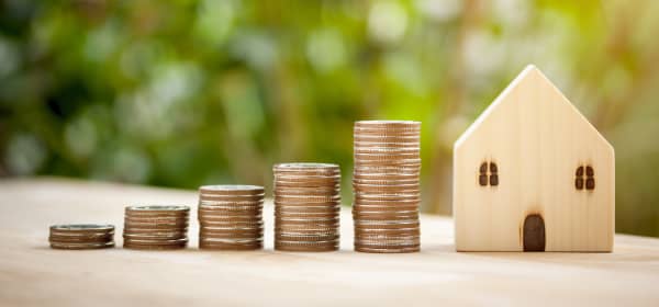 Münzstapel in verschiedenen Höhen neben kleinem Modellhaus aus Holz vor verschwommenem, grünen Hintergrund