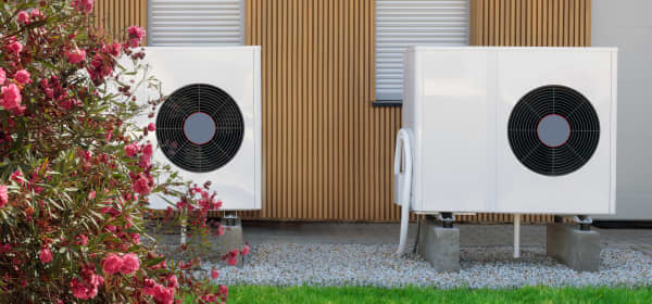 Zwei weiße Wärmepumpen vor einem Gebäude mit Holzfassade und mit Rollläden verschlossenen Fenstern