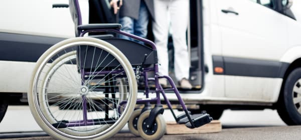 Rollstuhl steht vor einem weißen Van, aus dem ein Mann mit Gehstock aussteigt, wobei er von einer zweiten Person gestützt wird