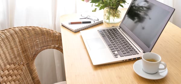 Tisch mit aufgeklapptem Laptop, Kaffeetasse, Notizblock und Blumenvase