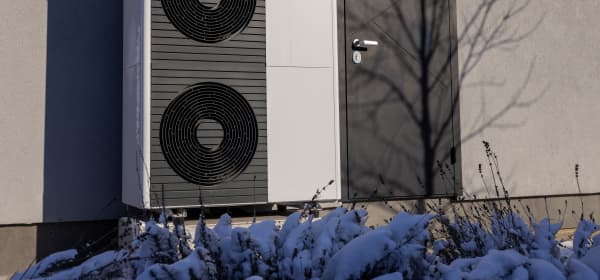 Luftwärmepumpen an der Fassade neben einer Eingangstür mit schneebedeckten Pflanzen im Vordergrund
