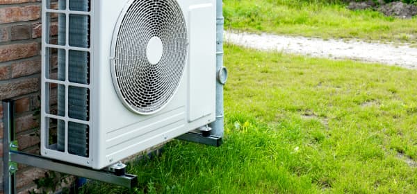 Luftwärmepumpe mit weißem Gehäuse an einer Hauswand mit Naturstein-Fassade mit grüner Wiese