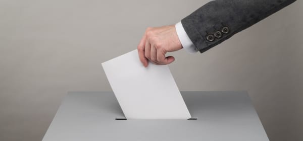 Mann mit Anzug steckt einen Zettel in eine Wahlurne, Aufnahme der Hand und des Arms