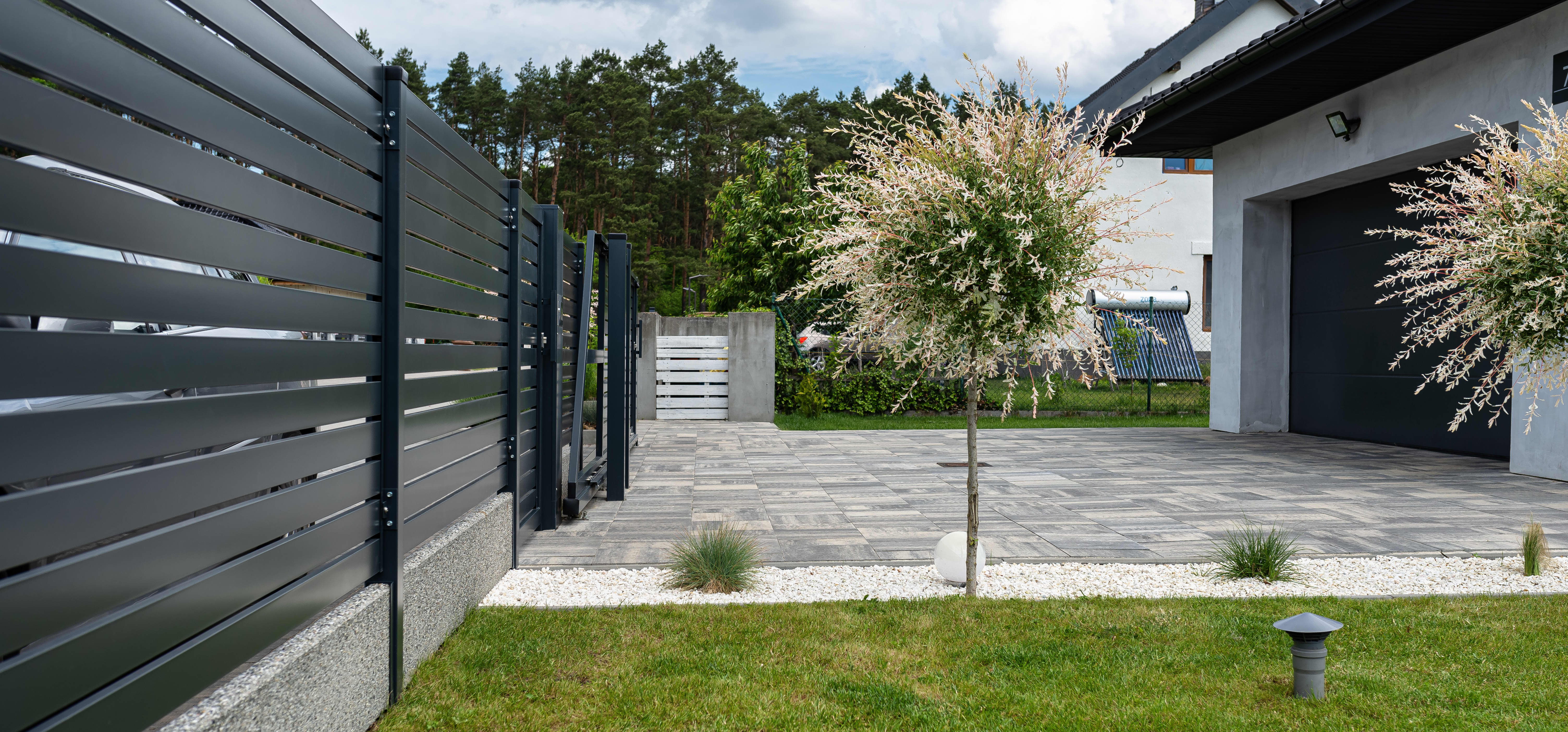 Modernes Wohnhaus mit gepflasterter Einfahrt und grüner Wiese. Das Grundstück wird durch einen hohen Zaun begrenzt.