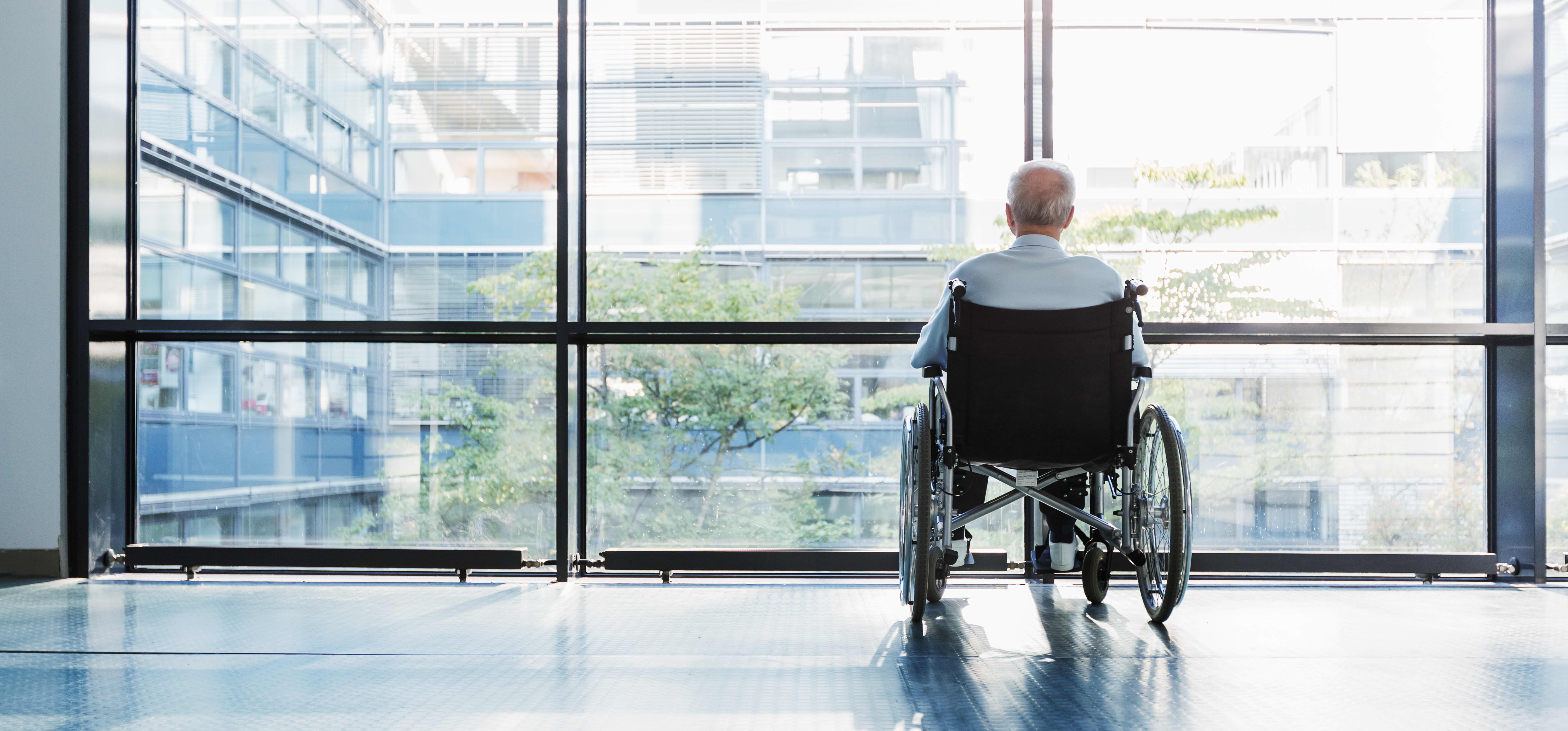 Senior im Rollstuhl blickt aus den bodentiefen Fenstern eines Gebäudes nach draußen