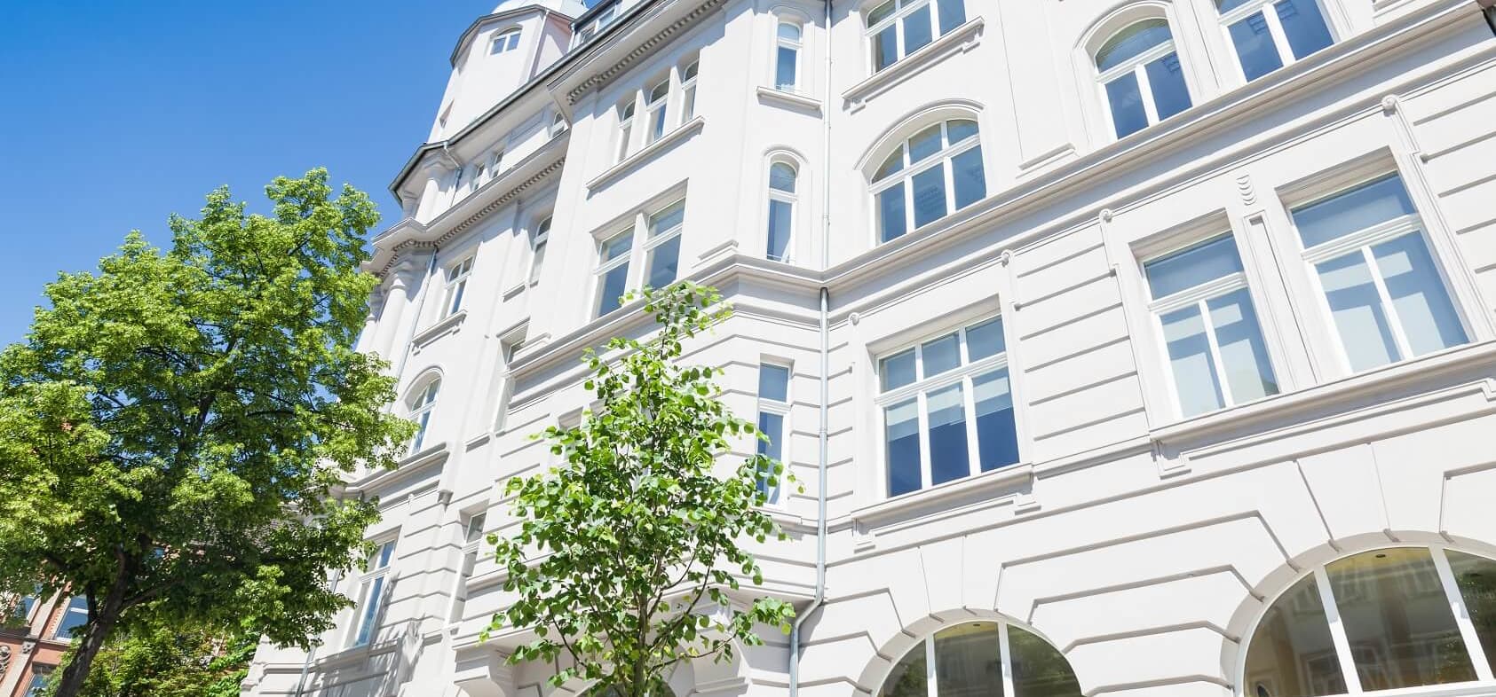 Saniertes Mehrfamilienhaus mit edler, weißer Fassade vor blauem Himmel mit grünen Bäumen davor