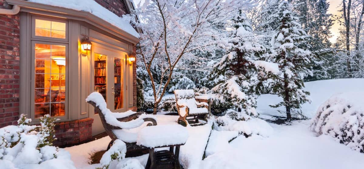 Schneebedeckter Garten und Terrasse, Blick in einen erleuchtetes Wohnzimmer eines Einfamilienhauses