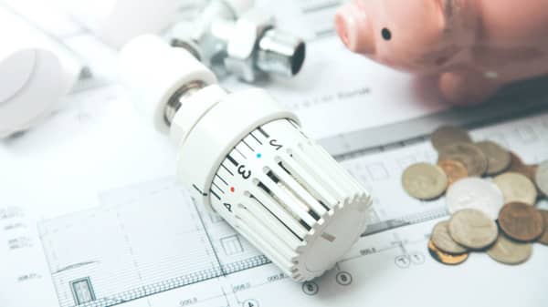 Thermostat einer Heizung liegt neben Kleingeld und Sparschwein auf Plänen.