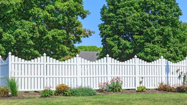 Hoher Zaun aus weißen Latten auf grüner Wiese mit grünen Laubbäumen im Hintergrund