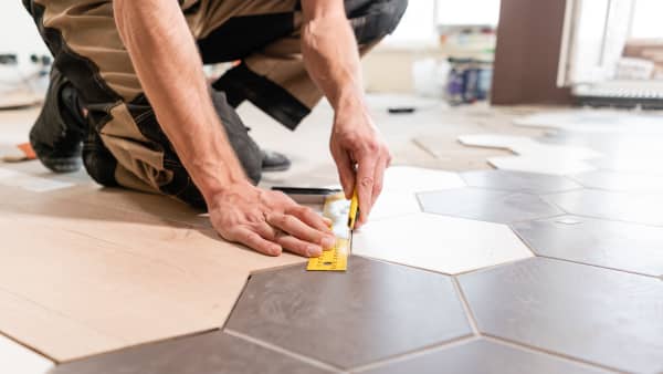 Mann in Arbeitskleidung kniet auf dem Boden und misst die Kante einer sechseckigen Bodenfliese zum Verlegen ab