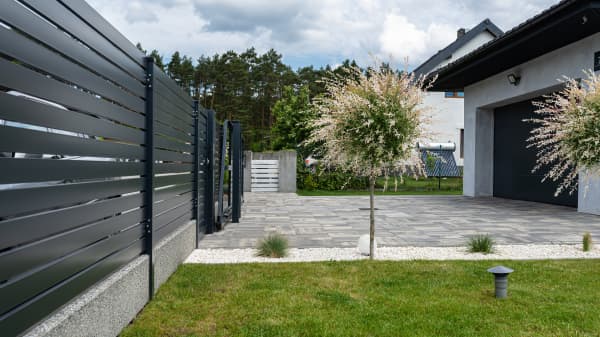 Modernes Wohnhaus mit gepflasterter Einfahrt und grüner Wiese. Das Grundstück wird durch einen hohen Zaun begrenzt.