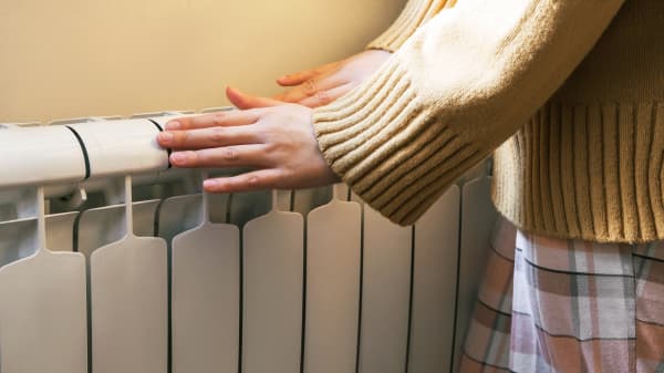 Frau in beigem Pullover und kariertem Rock wärmt ihre Hände an einem Heizkörper