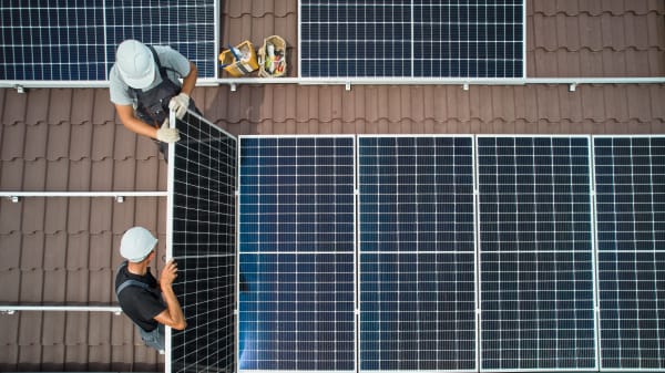 Ansicht von oben auf ein braun gedecktes Dach, auf dem zwei Arbeiter Solarmodule installieren