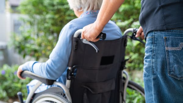 Rückansicht eines jüngeren Mannes, der eine Seniorin in einem Rollstuhl schiebt