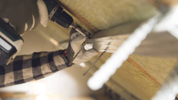 Nahaufnahme der Hände eines Arbeiters, der mit einem Akkuschrauber Bauteile innen an einer Dachschräge befestigt