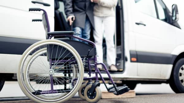 Rollstuhl steht vor einem weißen Van, aus dem ein Mann mit Gehstock aussteigt, wobei er von einer zweiten Person gestützt wird