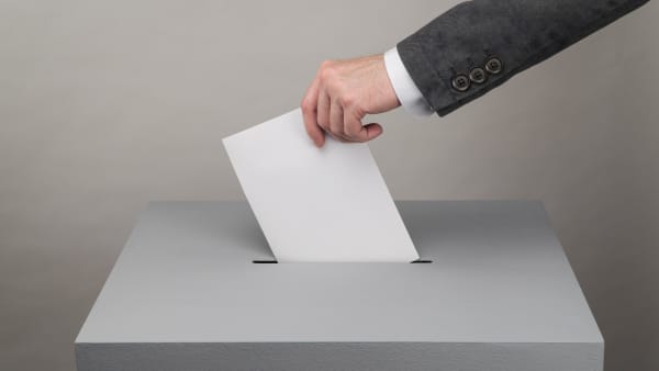 Mann mit Anzug steckt einen Zettel in eine Wahlurne, Aufnahme der Hand und des Arms