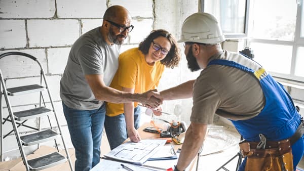 Ein Bauarbeiter mit Schutzhelm und blauer Latzhose schüttelt einem Mann in einem grauen T-Shirt die Hand, während eine Frau im gelben Oberteil daneben steht und lächelt. Sie befinden sich in einem halbfertigen Raum und stehen um einen Tisch mit ausgebreiteten Bauplänen und Werkzeugen, was auf eine Planung oder Besprechung hinweist.