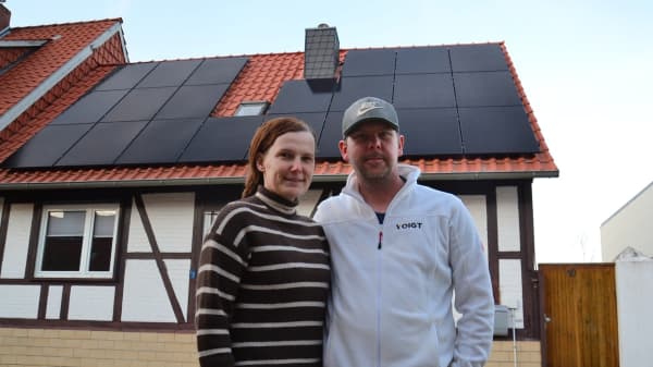 Eine Frau und ein Mann stehen vor einem Haus mit Solarmodulen auf dem Dach. Die Frau lächtelt und trägt einen gestreiften Pullover, der Mann eine weiße Jacke und eine Baseball Kappe. Sie präsentieren ihre neue Solaranlage.