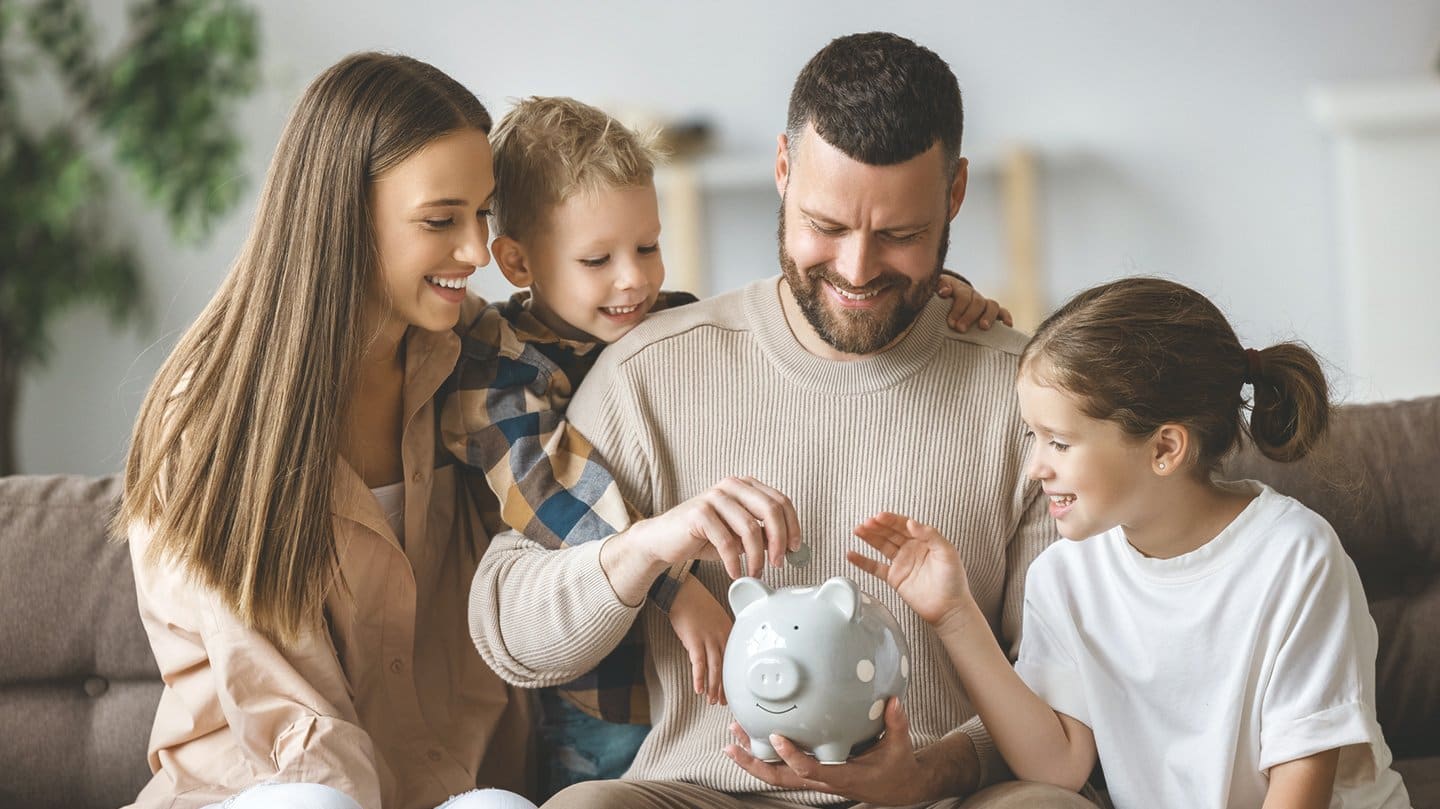 Mutter, Vater und zwei Kinder sitzen zu Hause auf dem Sofa und stecken fröhlich eine Münze ins Sparschwein.